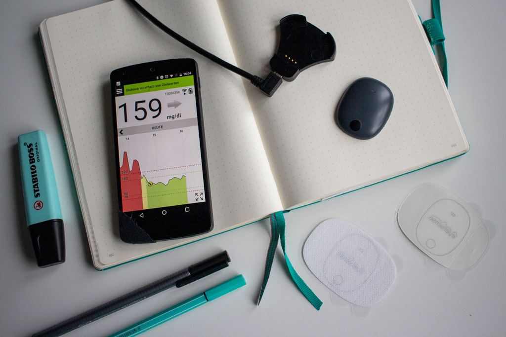 Auf einem aufgeschlagenem Notizbuch liegt der Tranmitter und das Ladegrät des Eversense XL, sowie ein Smartphone, auf dem man Glucosewerte sehen kann.